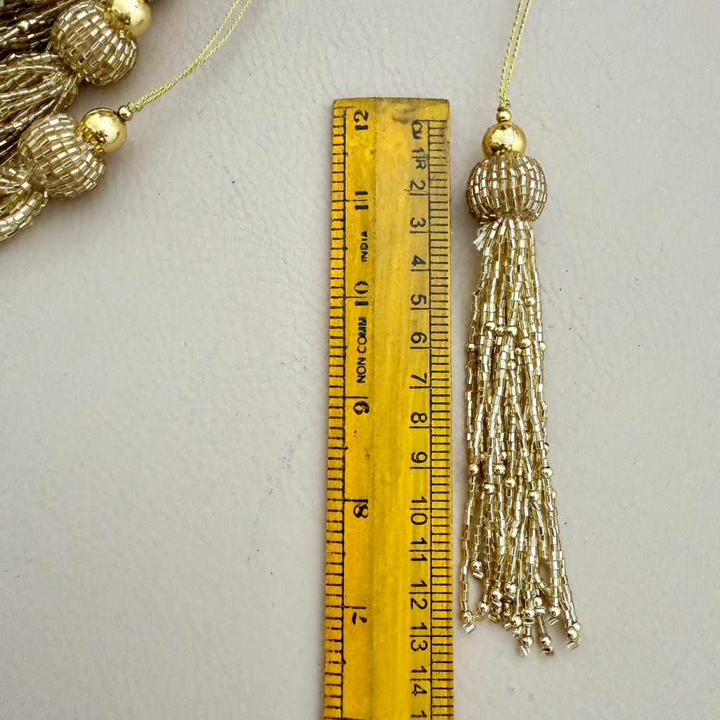 Gold tassel with decorative beads next to a ruler on a light gray background
