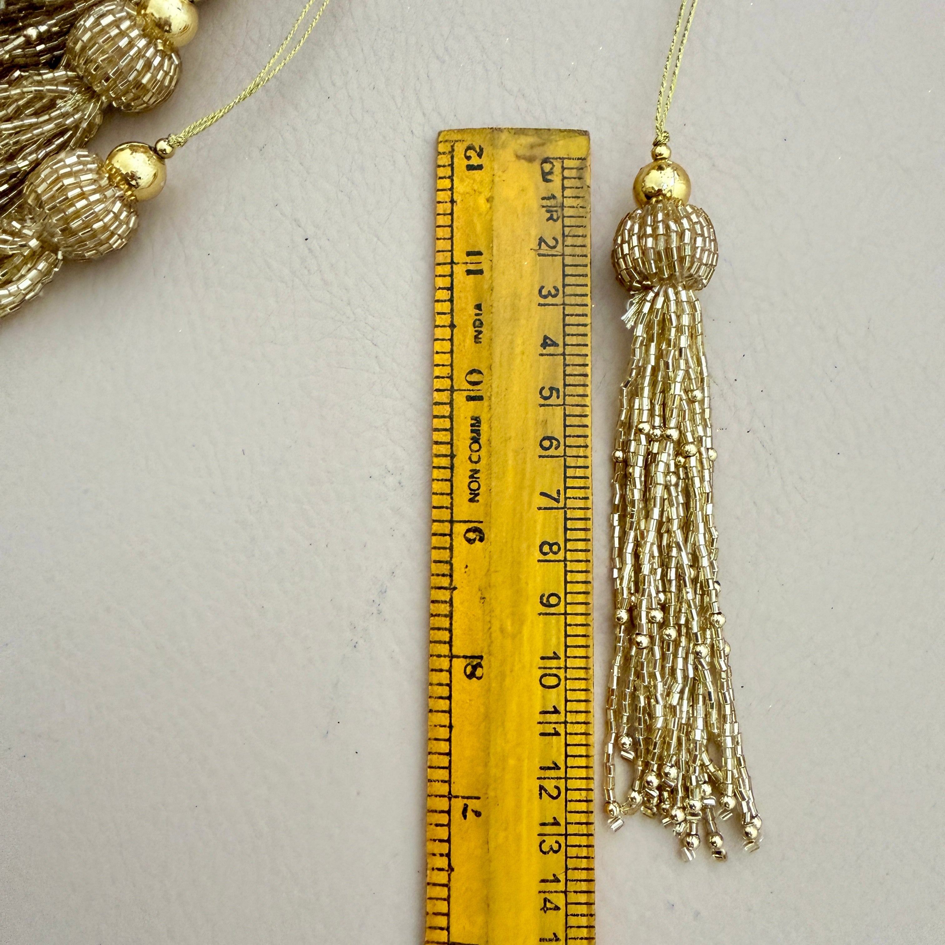 Gold tassel with decorative beads next to a ruler on a light gray background