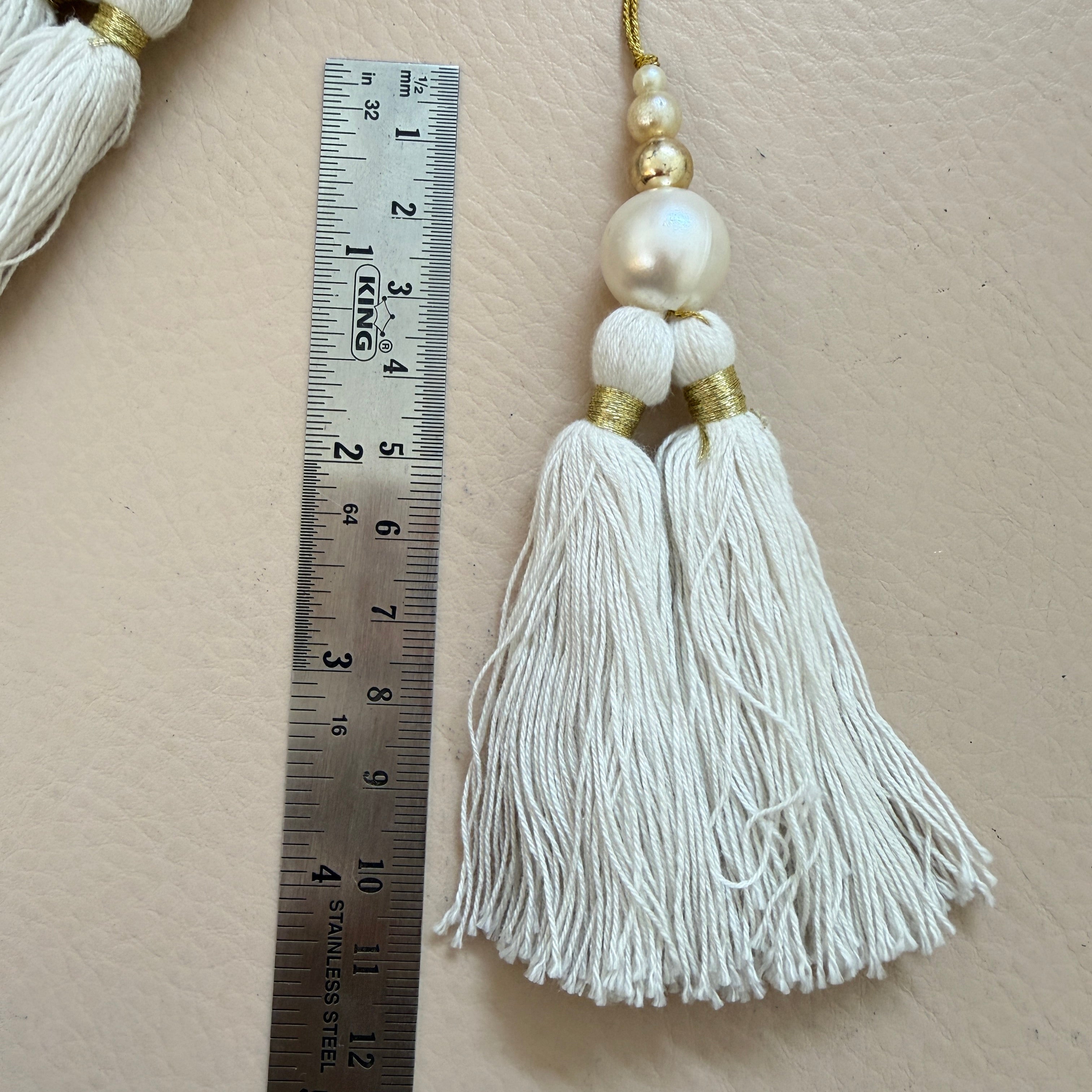 White tassel with gold accents and pearls next to a ruler on a beige background