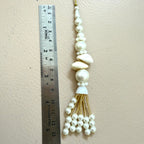 Decorative tassel with pearls and a shell on a beige background, next to a ruler for scale.