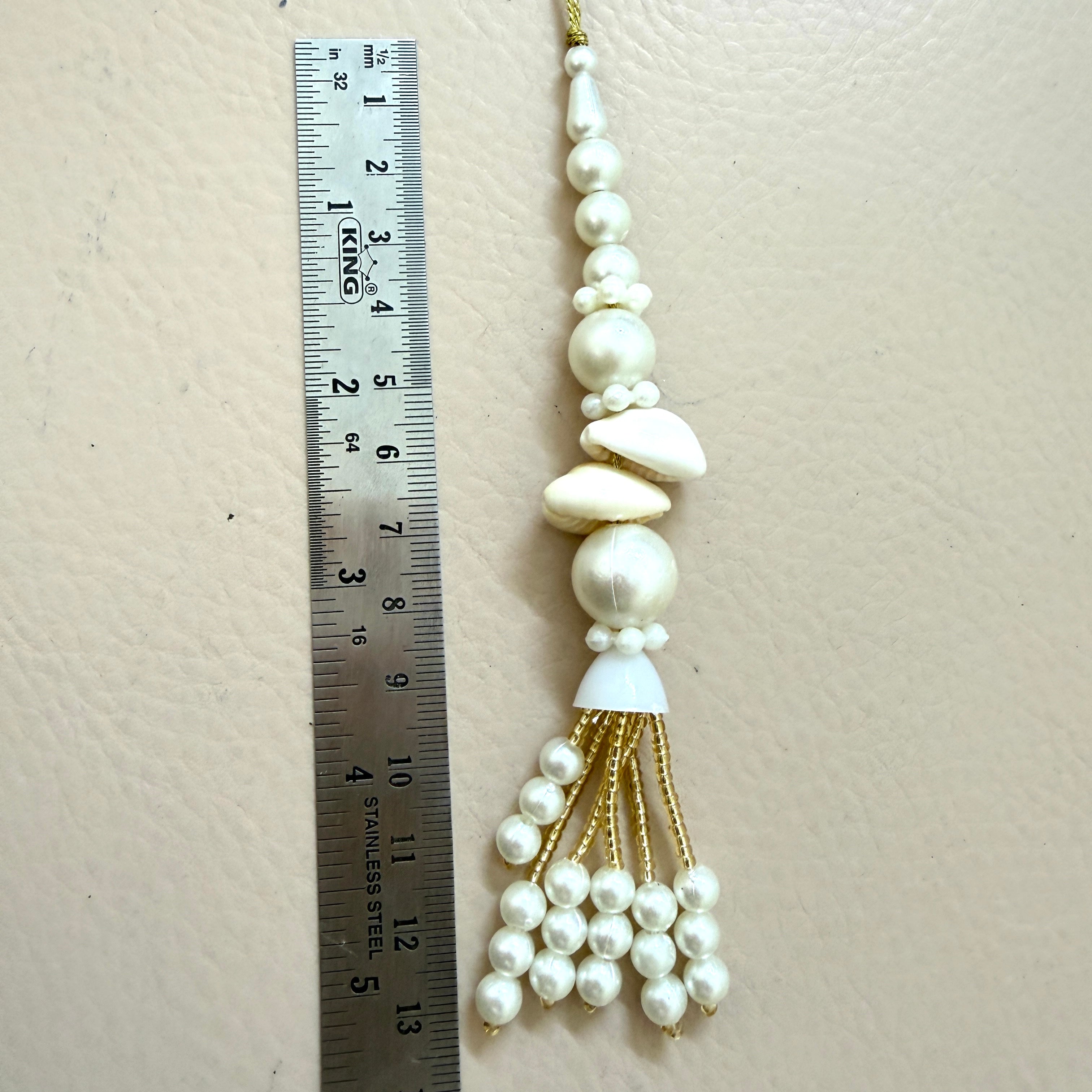 Decorative tassel with pearls and a shell on a beige background, next to a ruler for scale.