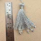 Silver tassel with beads next to a ruler on a beige background