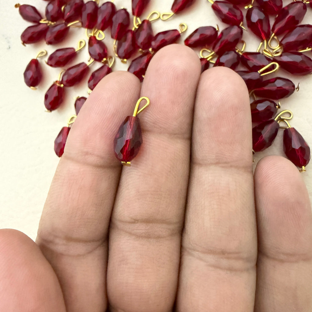Red teardrop-shaped jewelry pieces with gold hooks on a hand, with more pieces in the background.