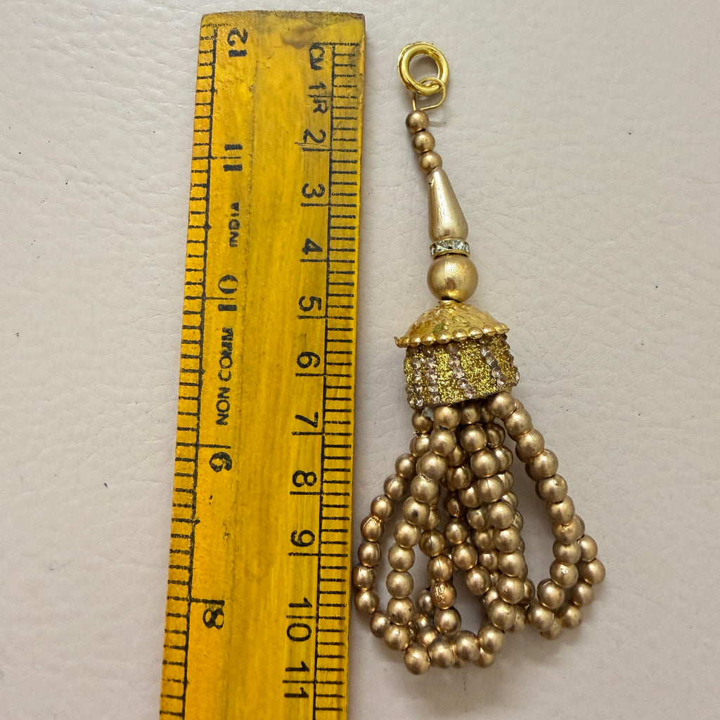 Gold tassel charm next to a ruler on a light gray background