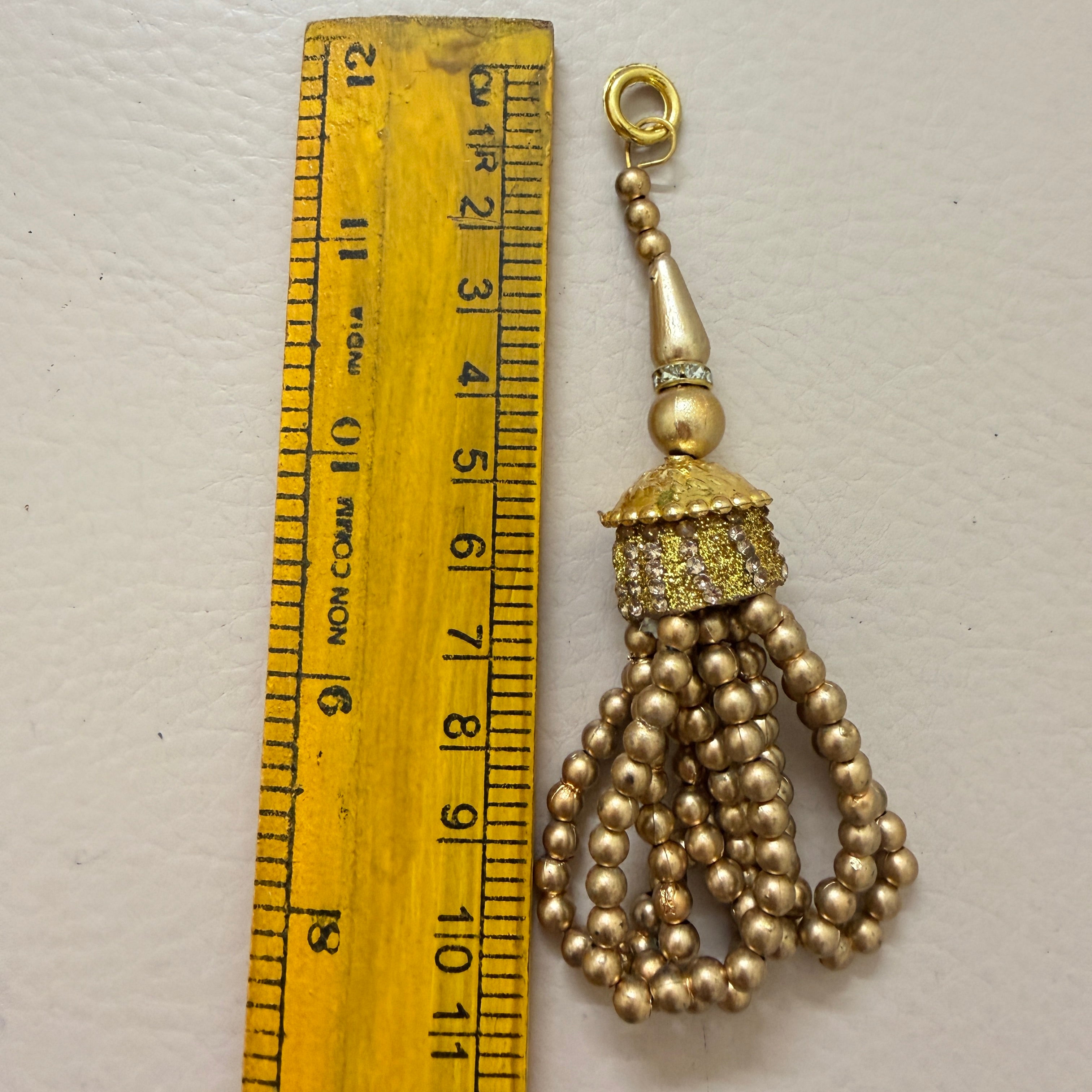 Gold tassel charm next to a ruler on a light gray background