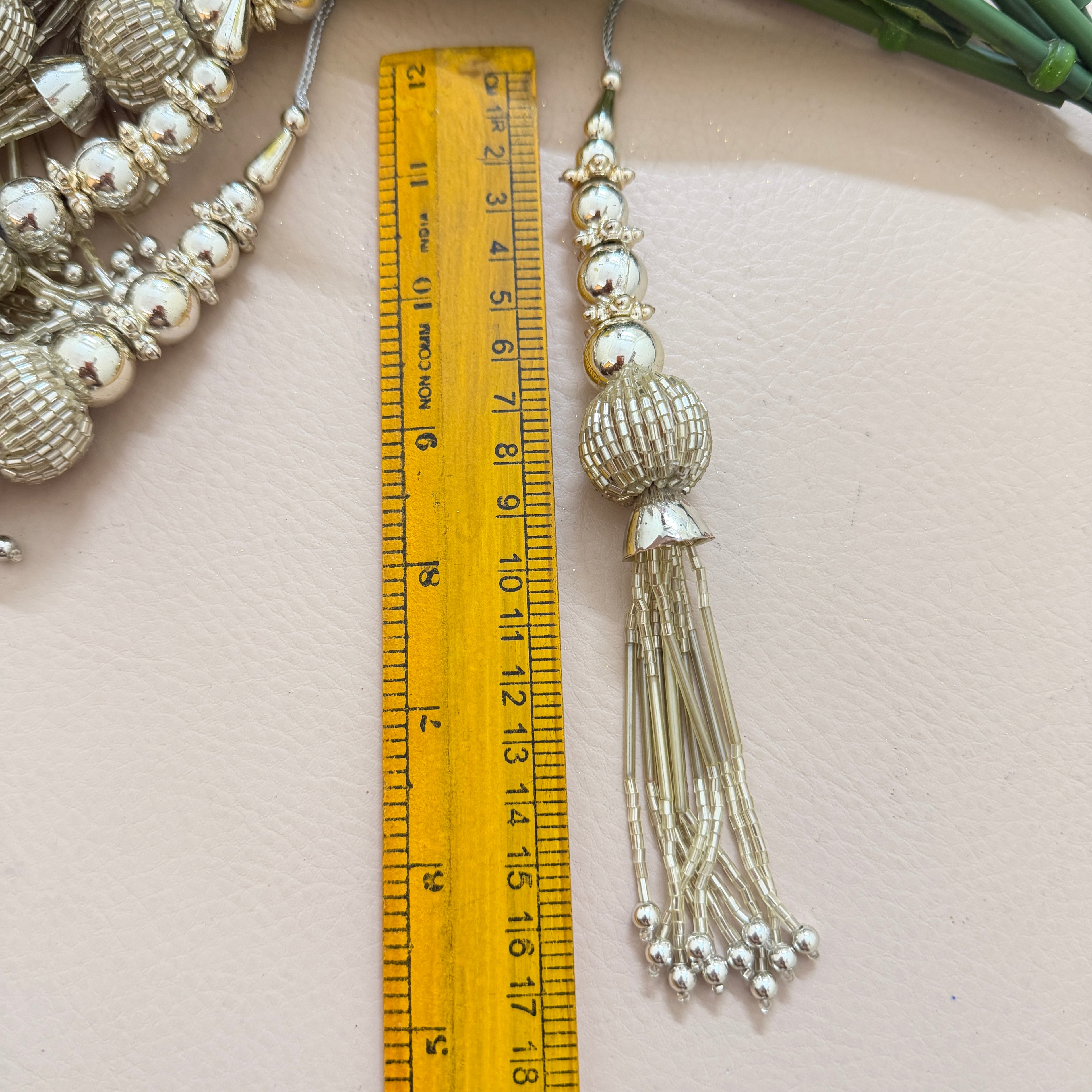 Decorative silver tassel with beads next to a ruler on a light pink background
