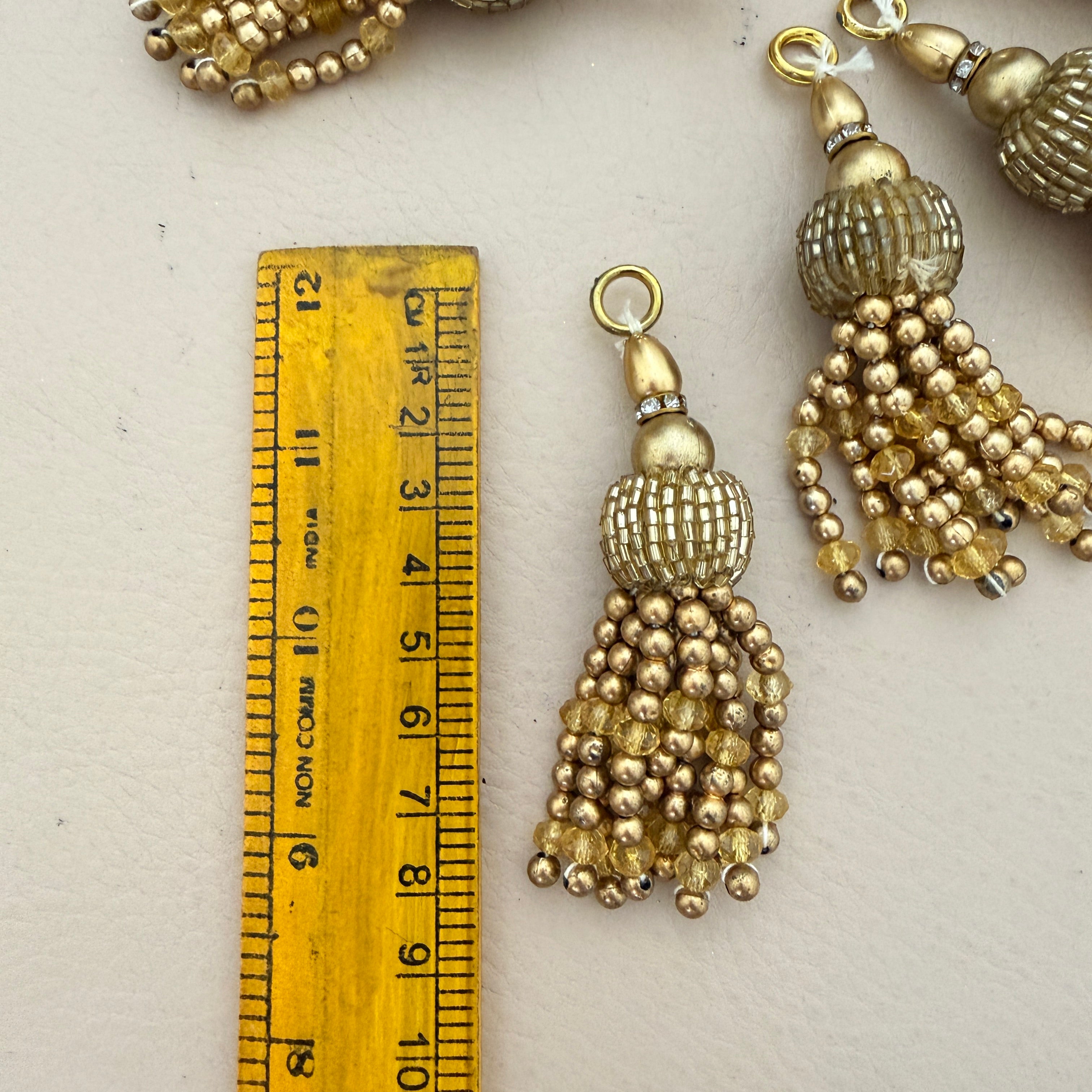 Gold tassel earrings with beads next to a wooden ruler for scale on a light background