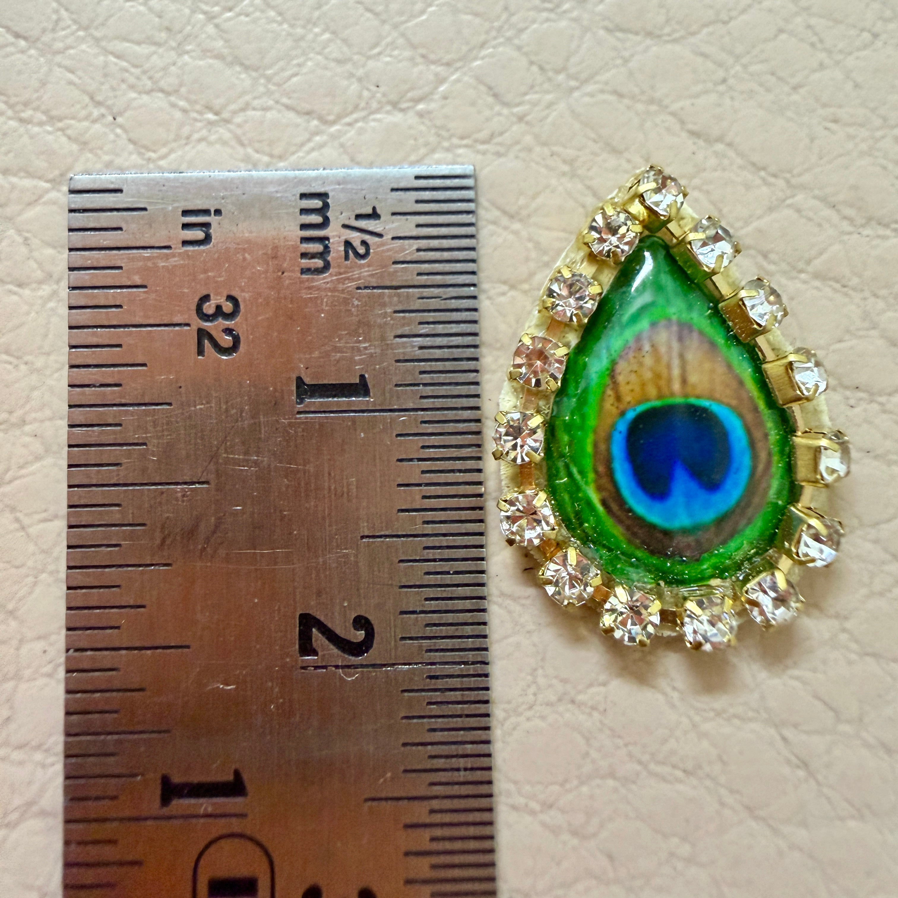 Teardrop-shaped peacock feather brooch with gemstones next to a ruler on a beige background