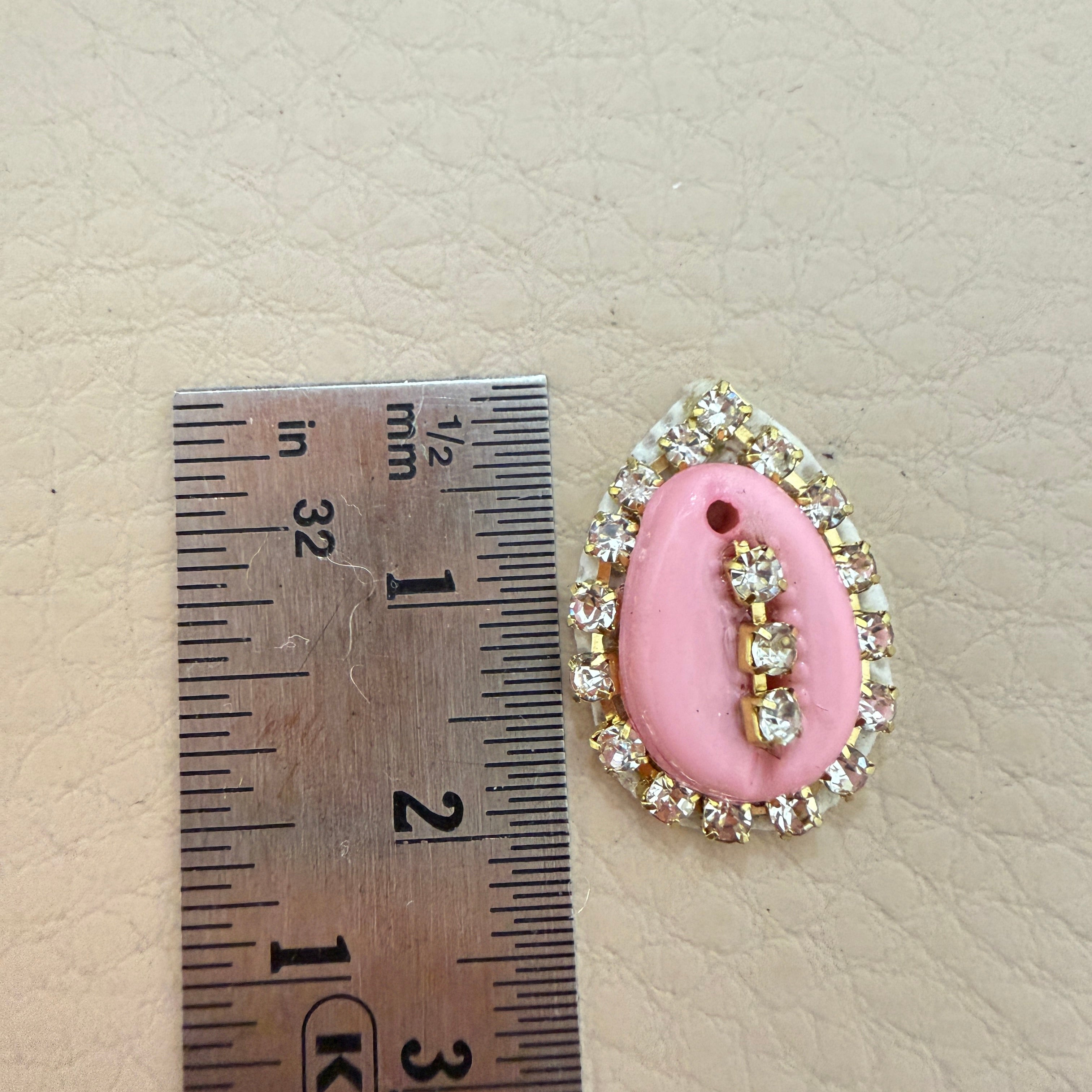 Pink oval pendant with decorative elements next to a ruler for scale on a beige background