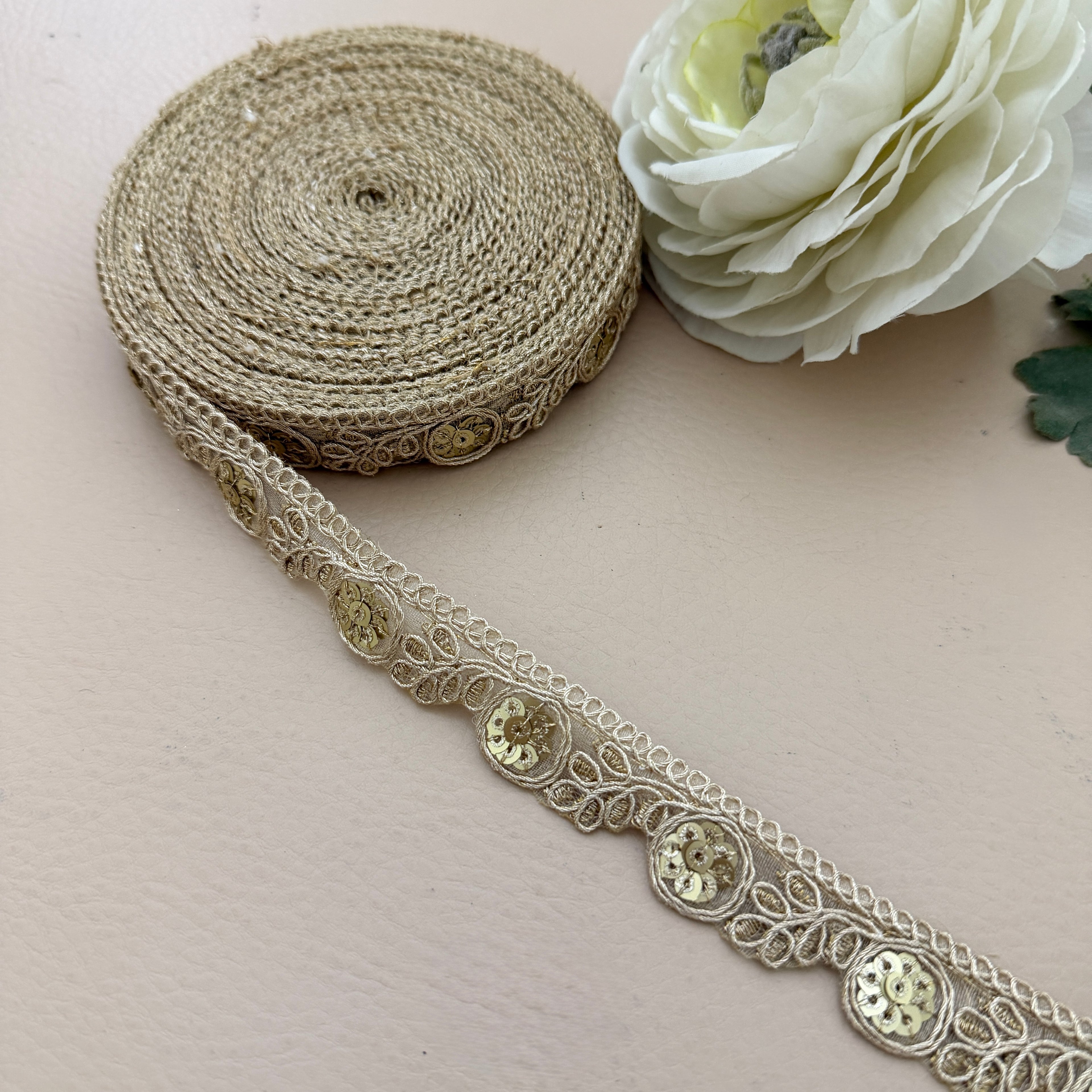 Decorative lace ribbon with gold embellishments on a light surface next to a white flower.