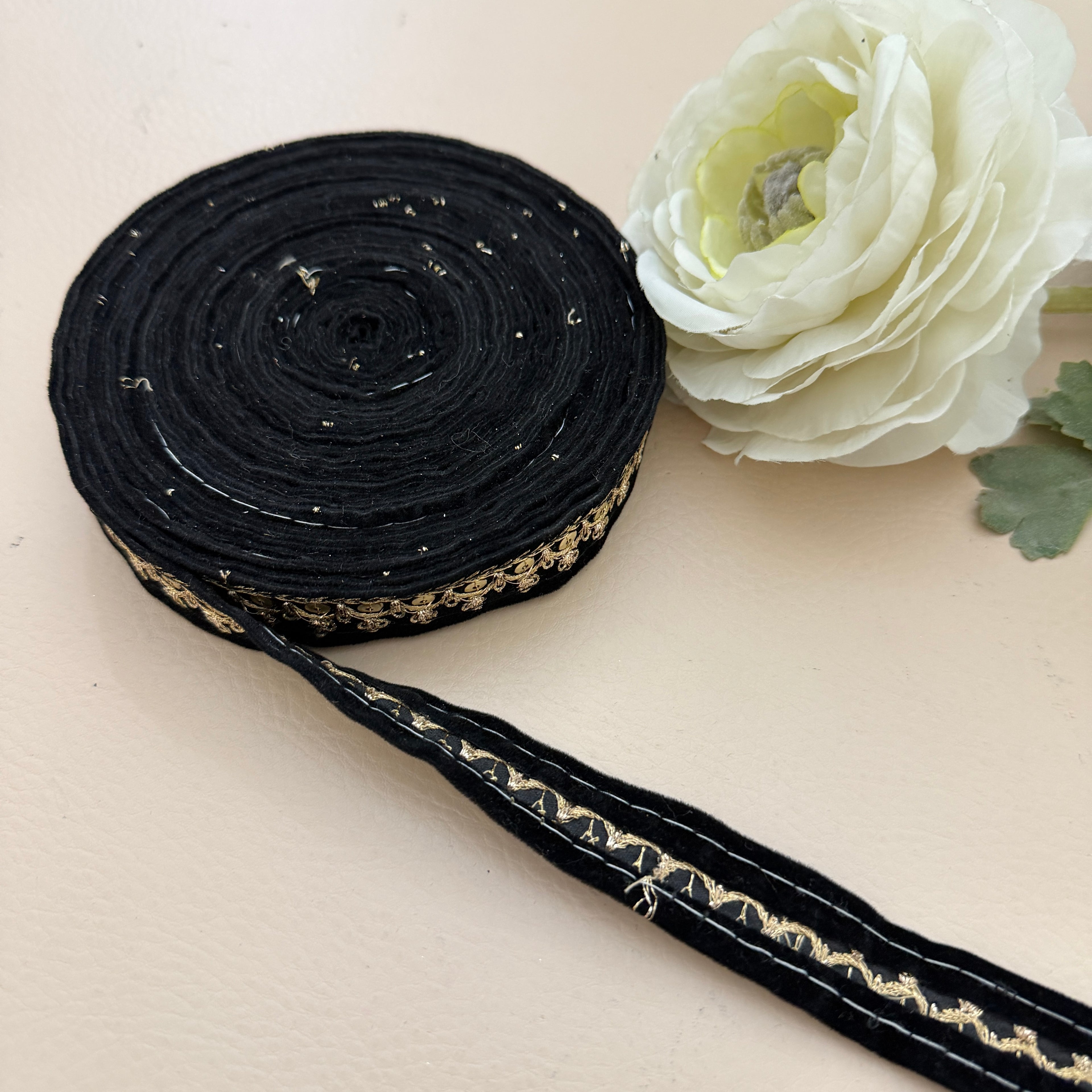 Black ribbon with gold embroidery on a beige surface next to a white flower.