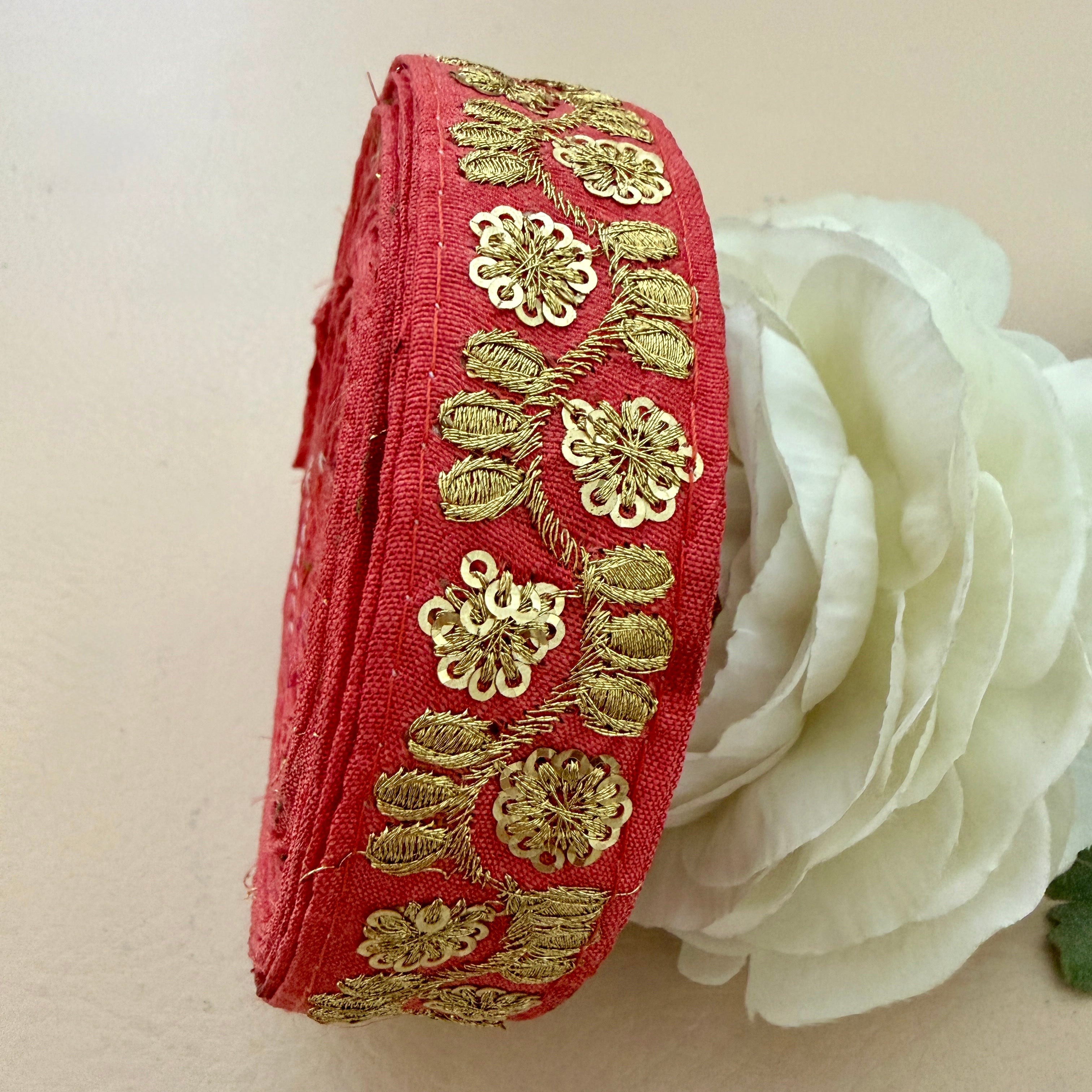 Red fabric ribbon with gold embroidery next to a white flower on a beige background