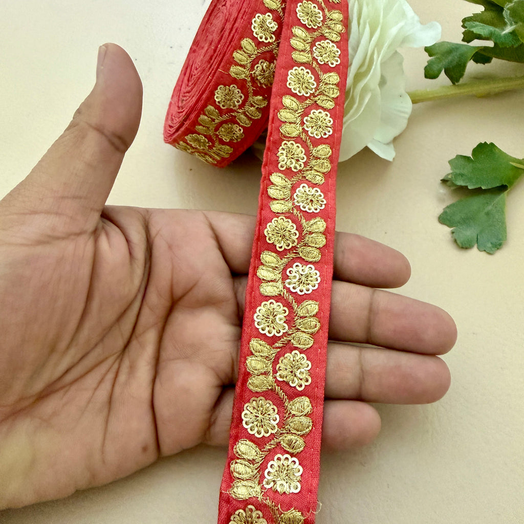 Red ribbon with gold floral embroidery held by a hand, with a roll of ribbon and flowers in the background.