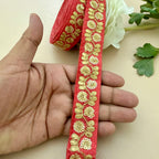 Red ribbon with gold floral embroidery held by a hand, with a roll of ribbon and flowers in the background.