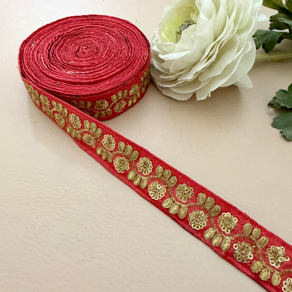 Red ribbon with gold floral embroidery next to a white flower on a beige background