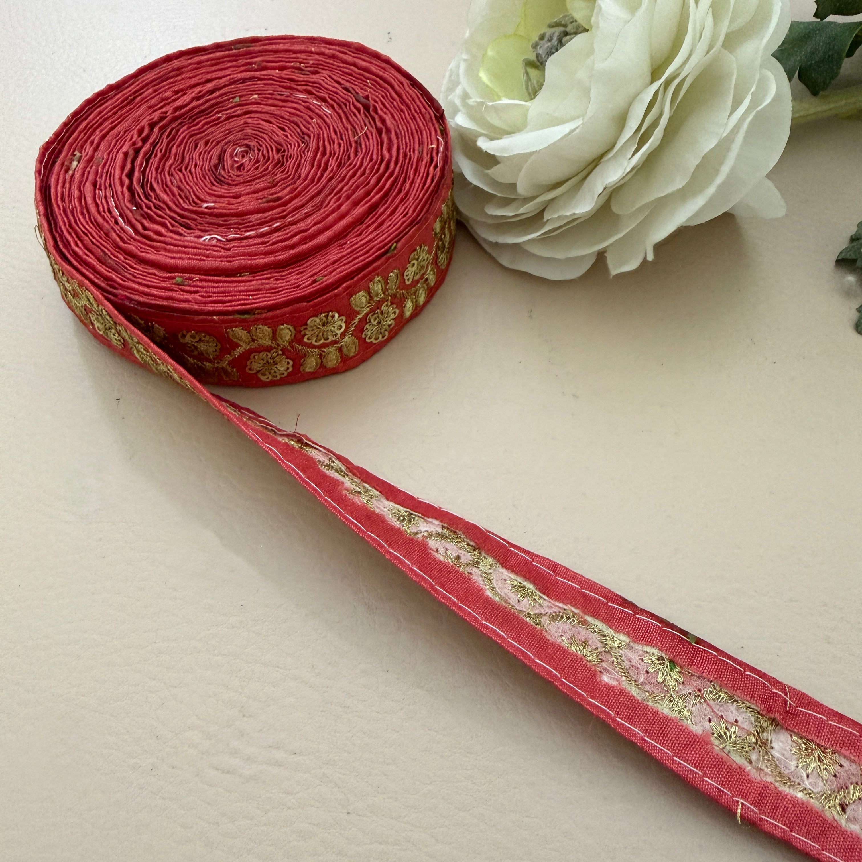 Roll of red fabric with gold embroidery next to a white flower on a beige background