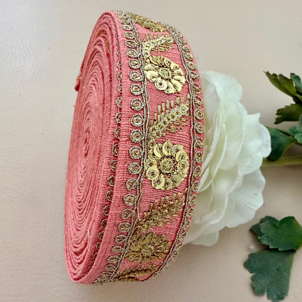 Roll of pink fabric with gold embroidery next to a white flower on a beige background