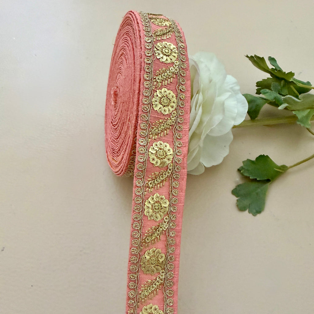 Decorative pink ribbon with gold embroidery on a light background with a white flower and green leaves.