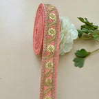 Decorative pink ribbon with gold embroidery on a light background with a white flower and green leaves.