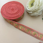 Roll of pink ribbon with gold floral patterns next to a white flower on a beige surface
