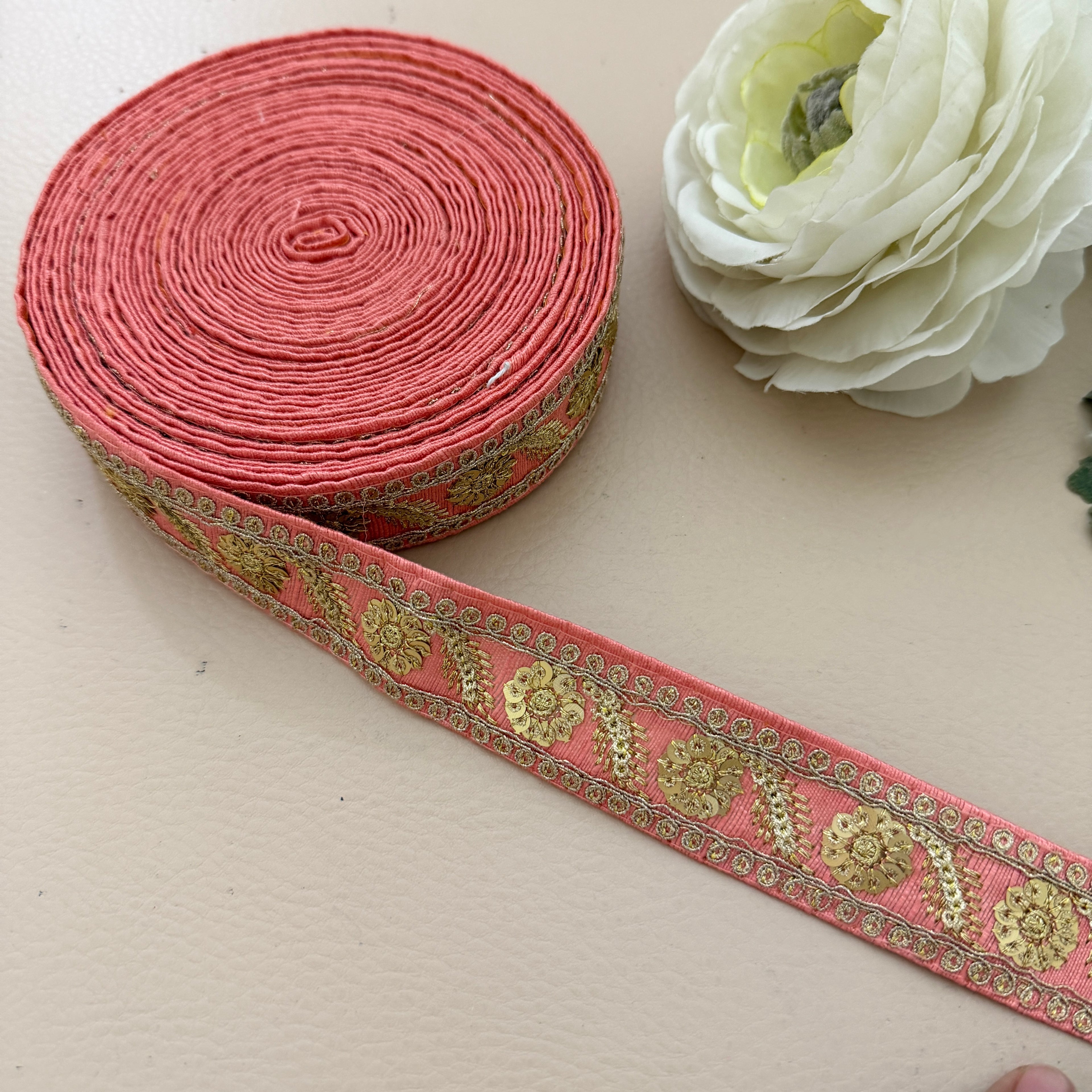 Roll of pink ribbon with gold floral patterns next to a white flower on a beige surface