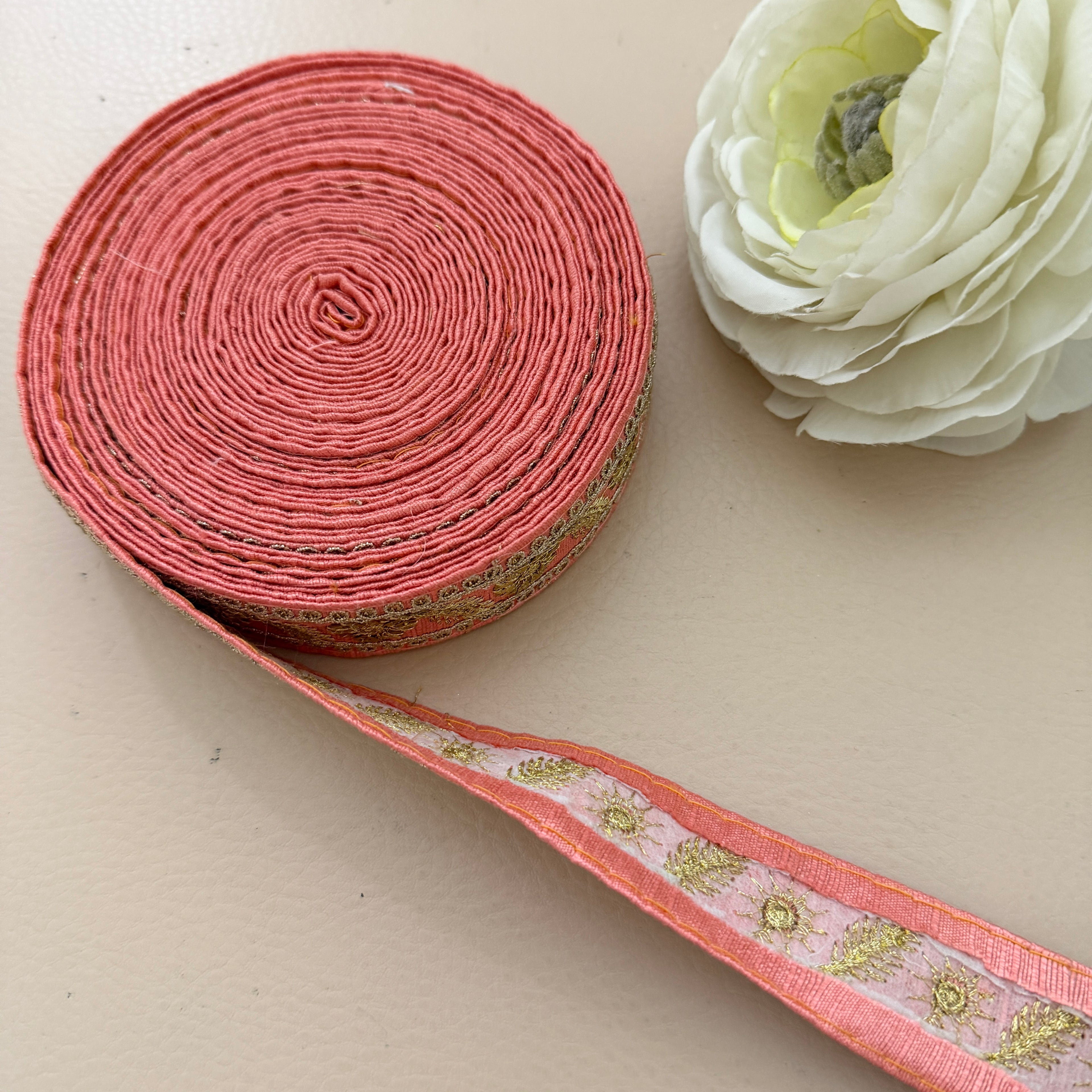 Roll of pink ribbon with decorative pattern next to a white flower on a beige background