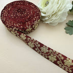 Roll of maroon ribbon with gold floral patterns next to a white flower on a beige background