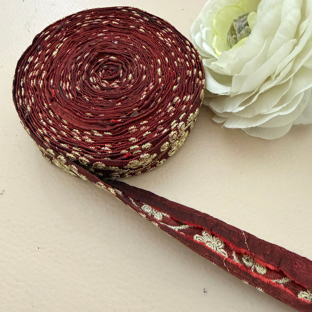 Roll of maroon ribbon with gold pattern next to a white flower on a light background