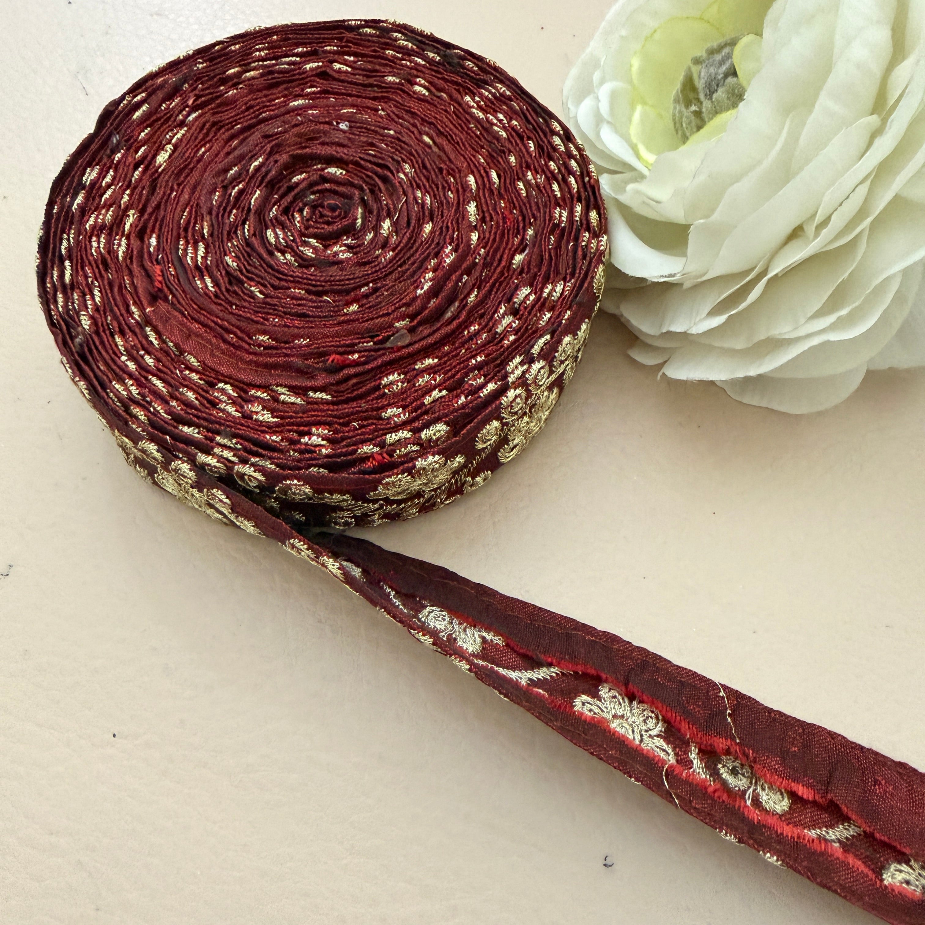 Roll of maroon ribbon with gold pattern next to a white flower on a light background