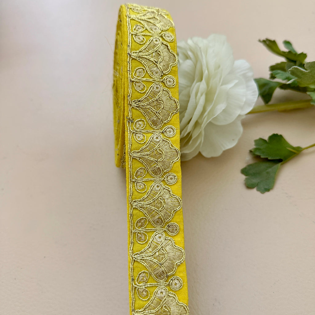 Yellow decorative ribbon with intricate patterns on a beige background with a white flower and green leaves.