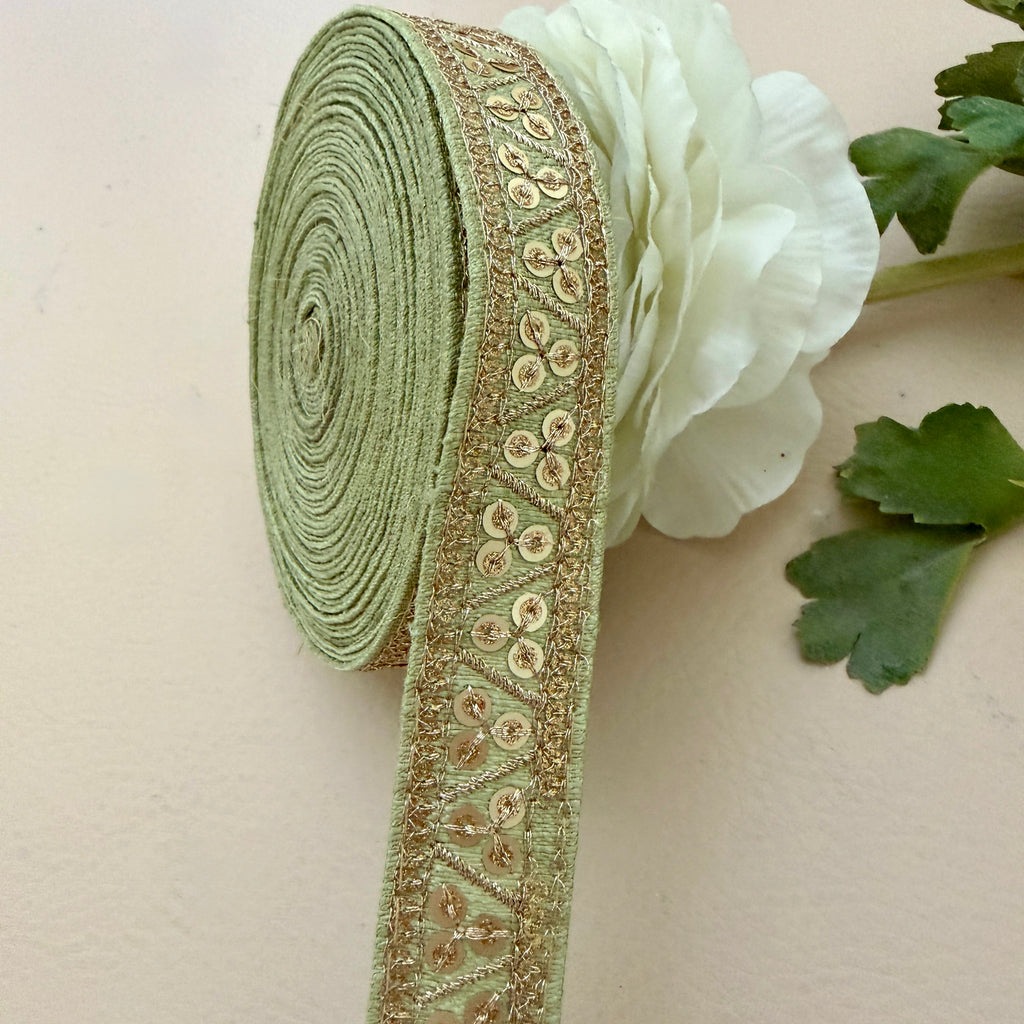 Green ribbon with gold embroidery next to a white flower on a beige background