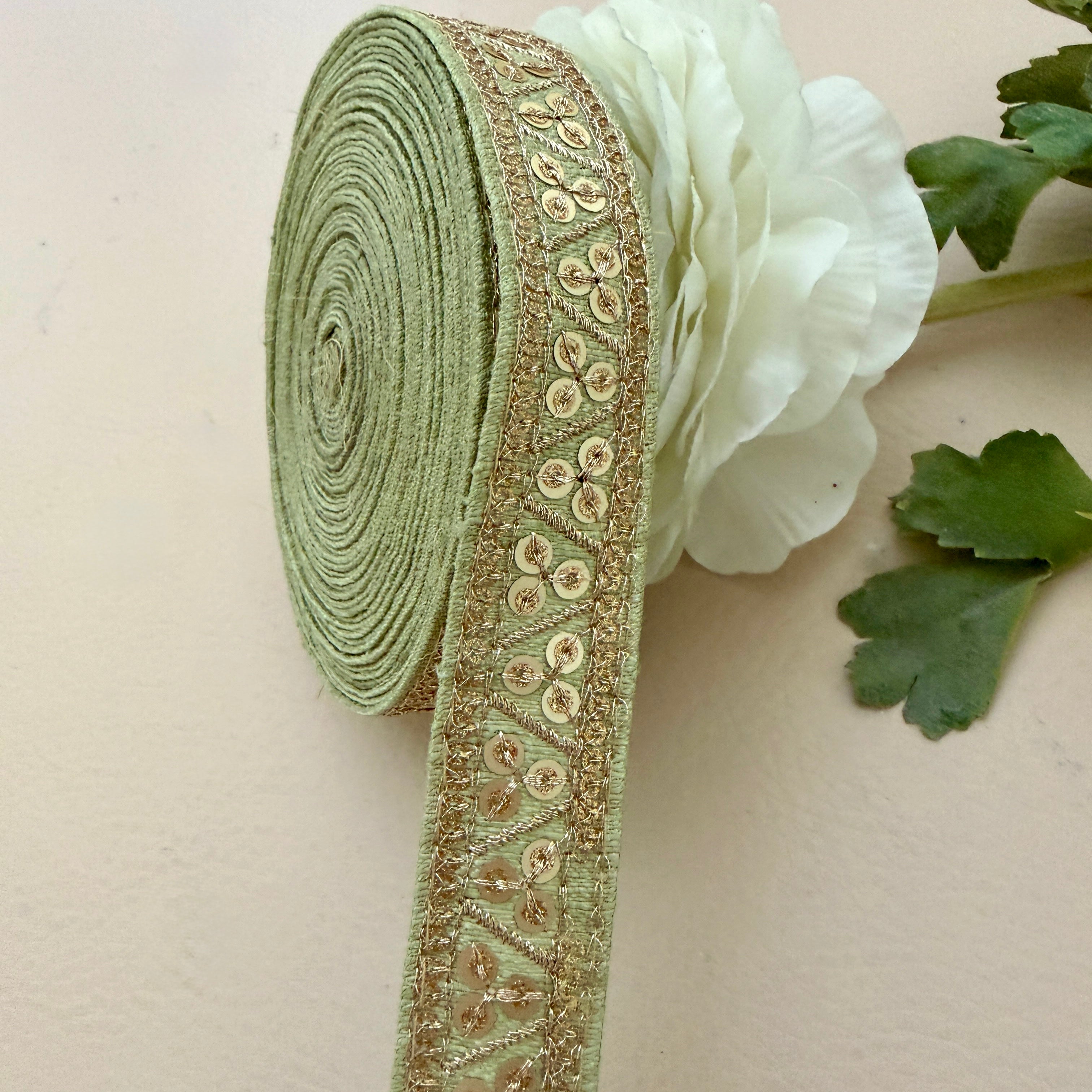Green ribbon with gold embroidery next to a white flower on a beige background