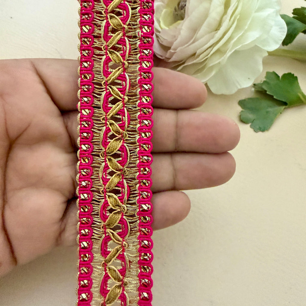 Decorative pink and gold trim held between fingers with a white flower in the background