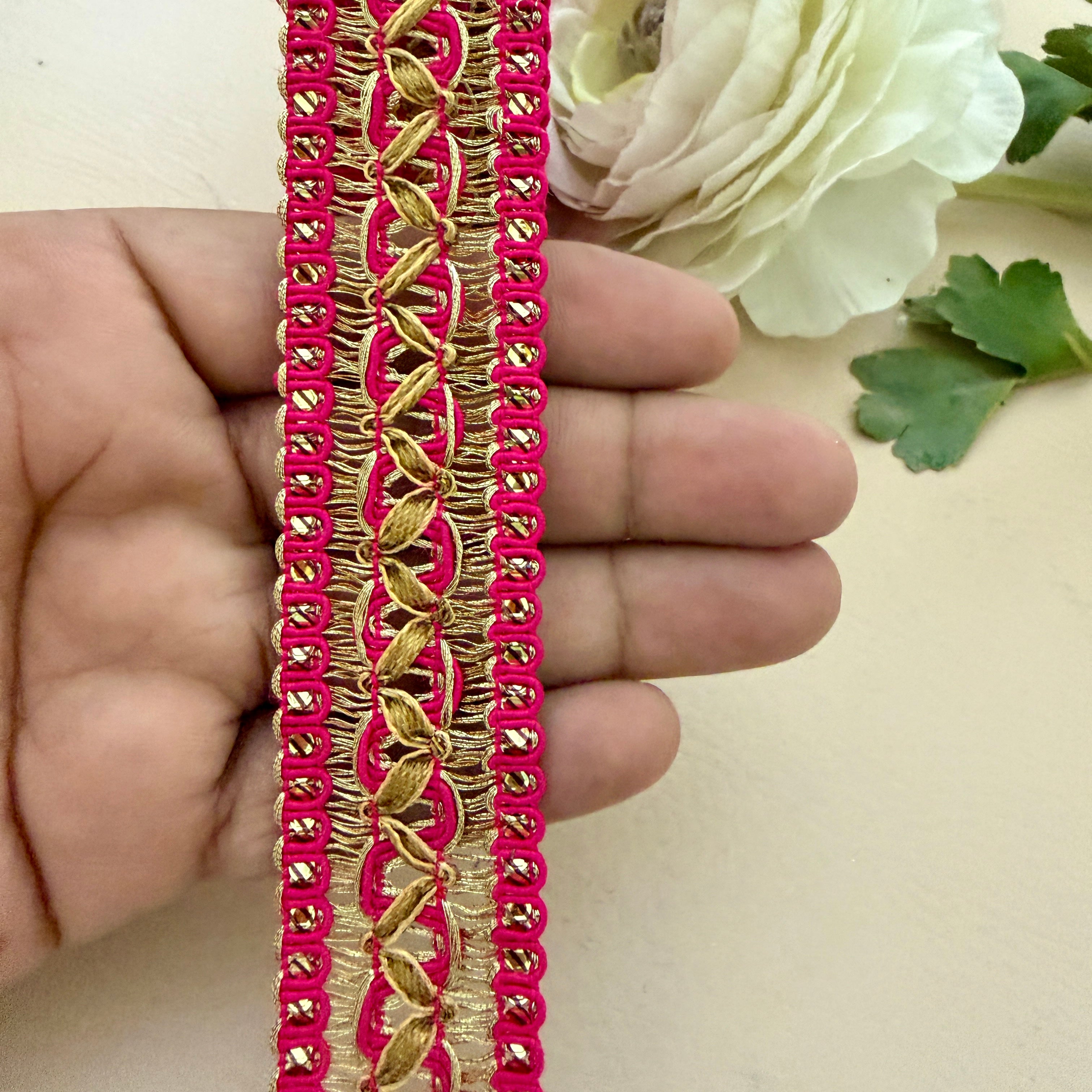 Decorative pink and gold trim held between fingers with a white flower in the background