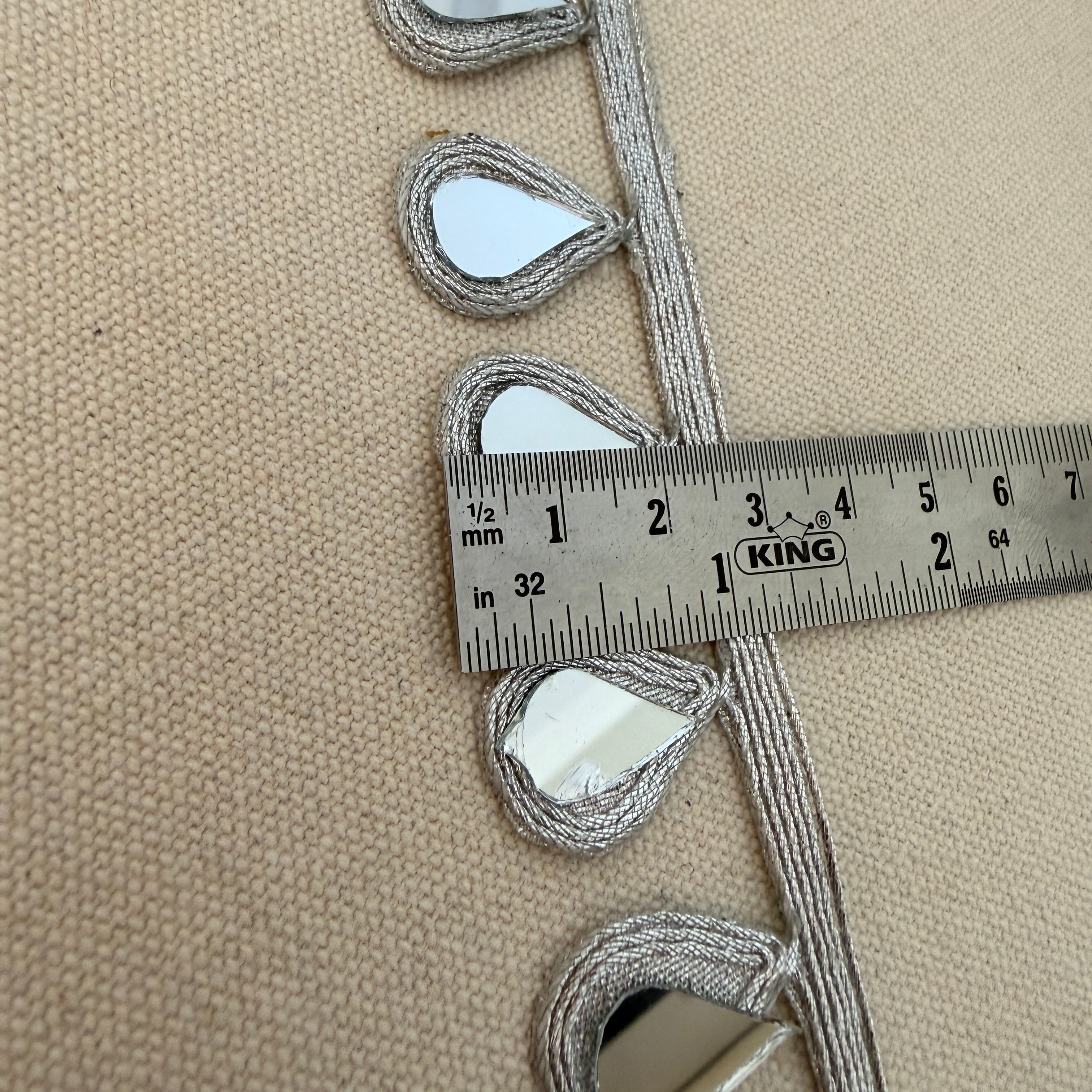 Decorative trim with teardrop shapes on a beige fabric background, measured with a ruler.