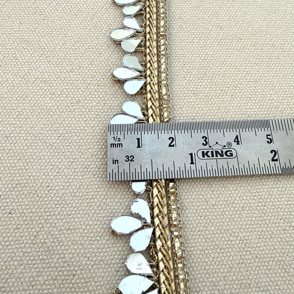 Decorative trim with white embellishments and gold accents being measured with a King ruler on a beige fabric background.