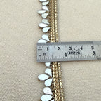 Decorative trim with white embellishments and gold accents being measured with a King ruler on a beige fabric background.