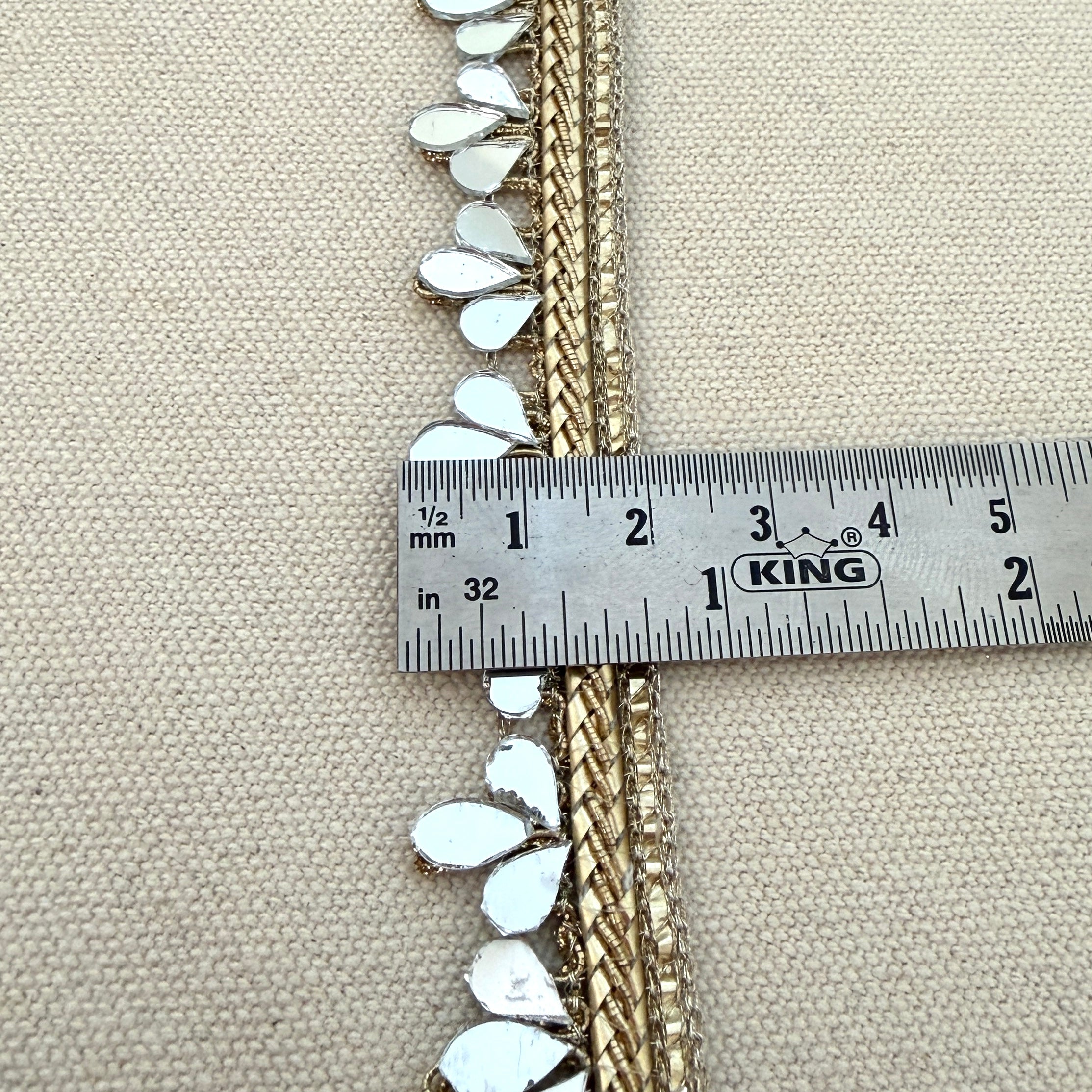 Decorative trim with white embellishments and gold accents being measured with a King ruler on a beige fabric background.
