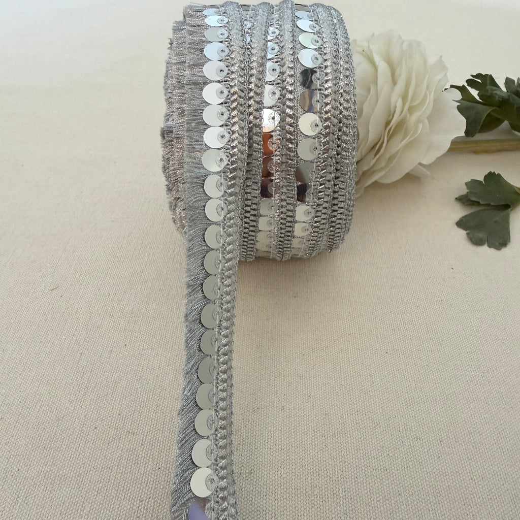 Roll of silver sequin ribbon on a beige fabric background with a white flower.