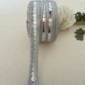 Roll of silver sequin ribbon on a beige fabric background with a white flower.