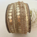 Close-up of a spool of decorative ribbon with gold sequins and fringe on a light background.