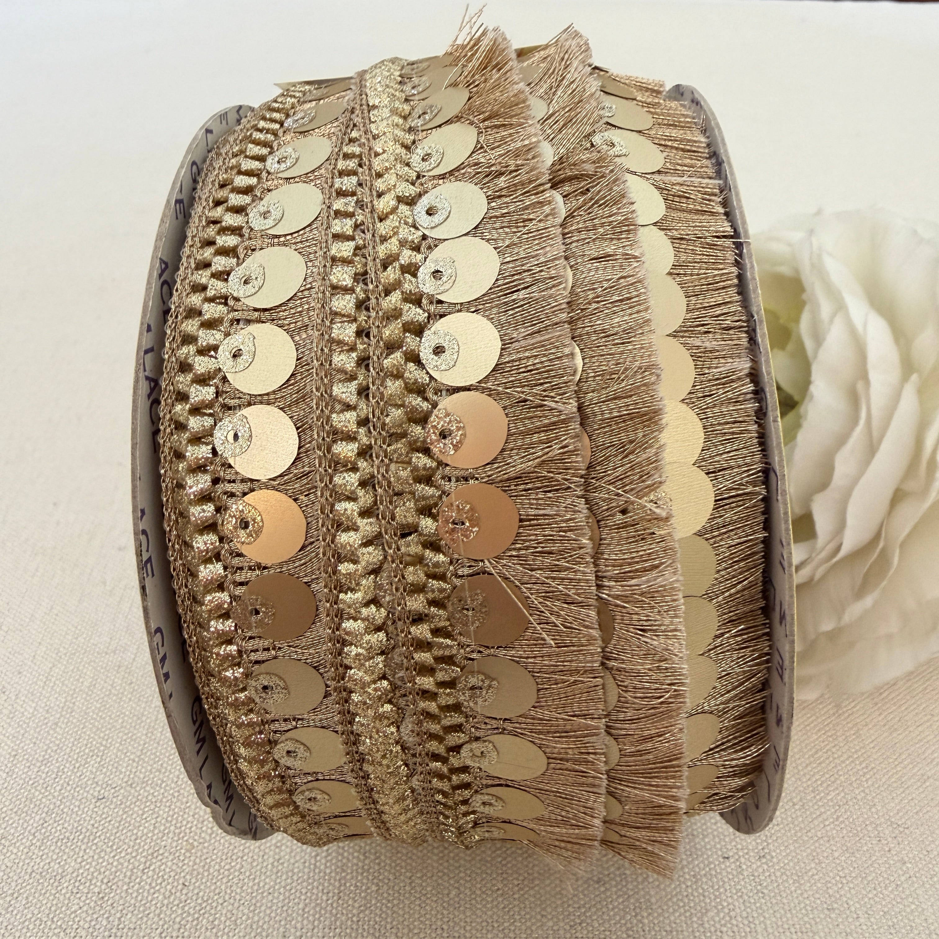 Close-up of a spool of decorative ribbon with gold sequins and fringe on a light background.