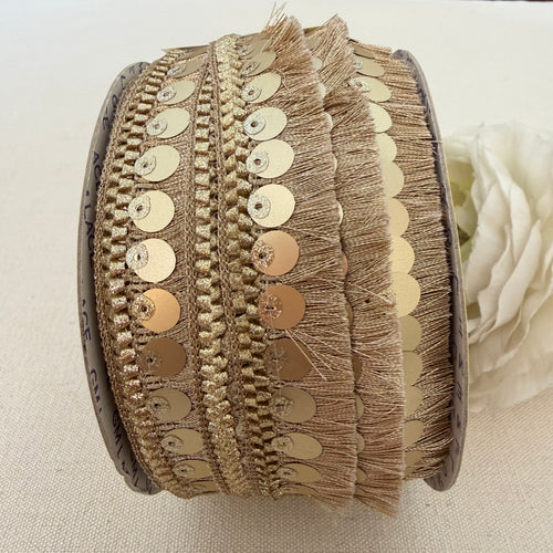 Close-up of a spool of decorative ribbon with gold sequins and fringe on a light background.