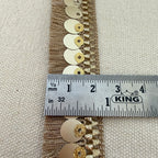 Decorative trim with gold and silver embellishments measured against a ruler on a beige fabric background.