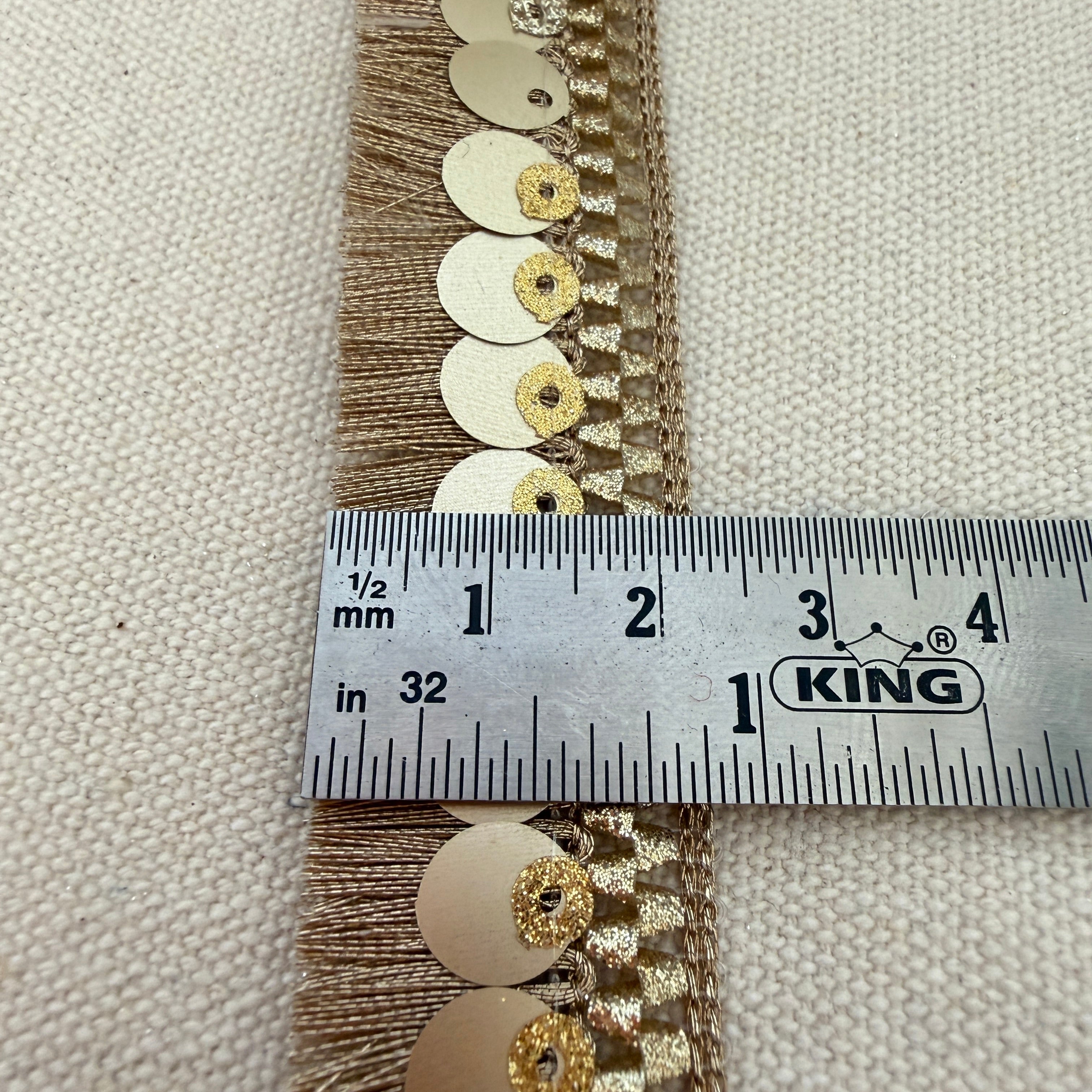 Decorative trim with gold and silver embellishments measured against a ruler on a beige fabric background.