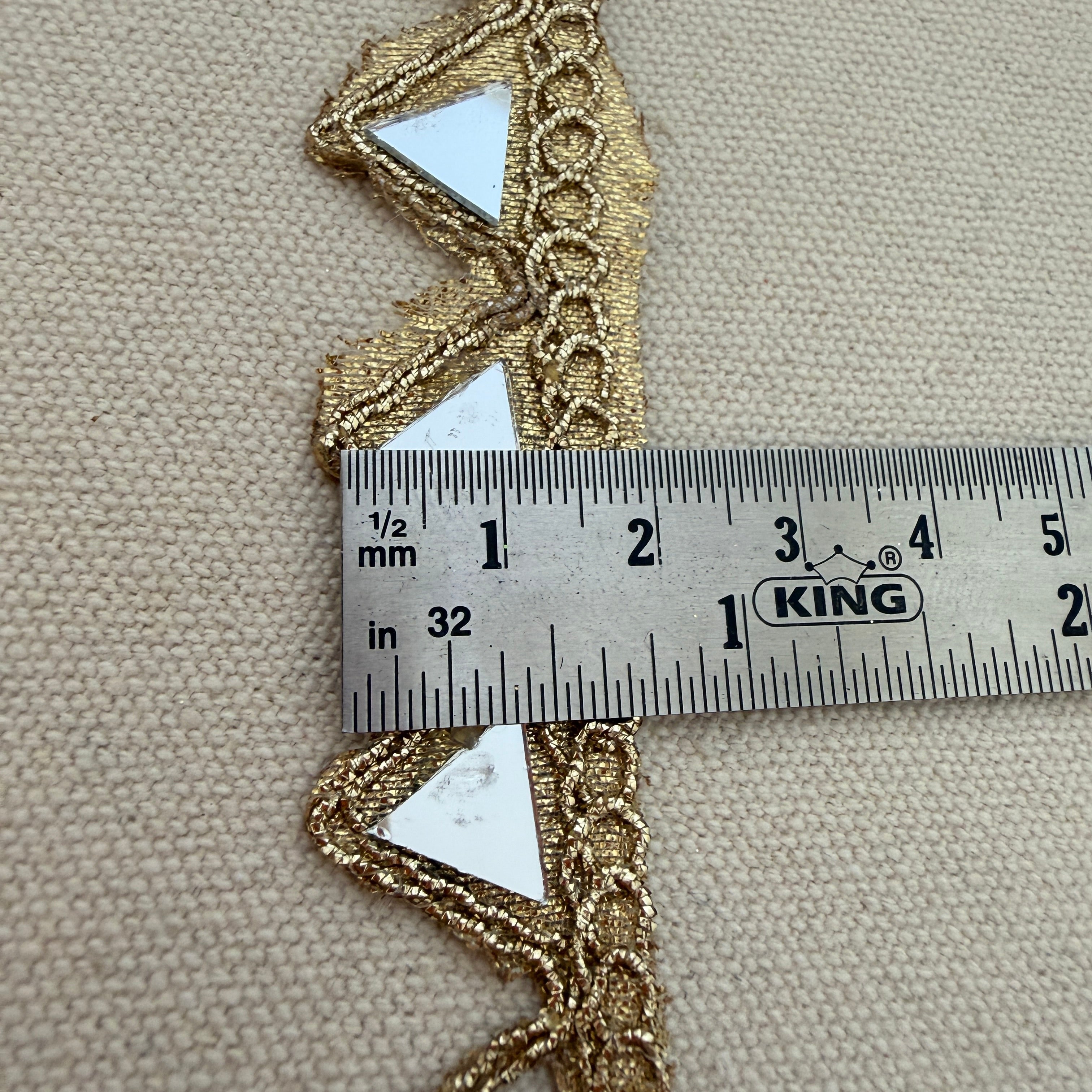 Gold lace trim with a ruler for scale on a beige fabric background