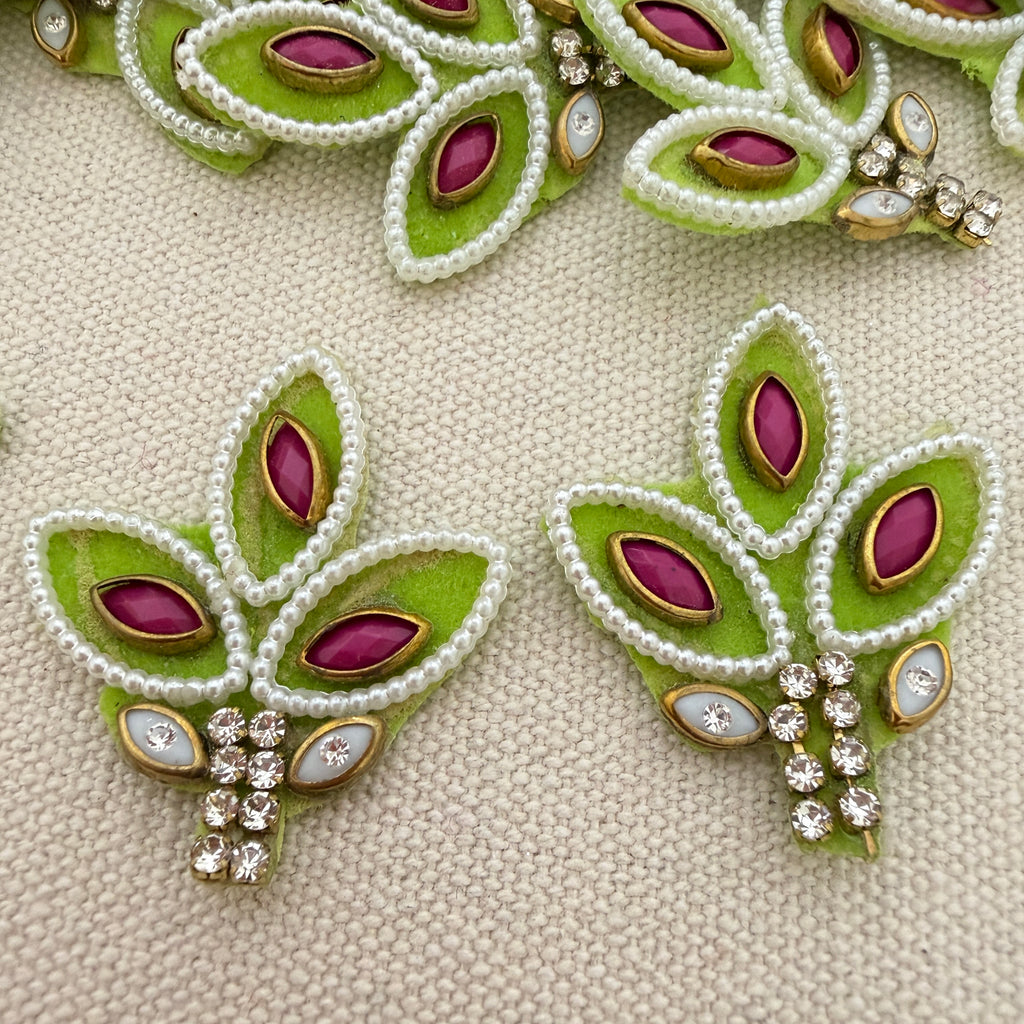 Decorative earrings with green leaves, purple stones, and white beads on a beige fabric background.