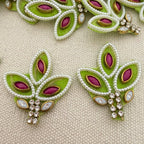 Decorative earrings with green leaves, purple stones, and white beads on a beige fabric background.