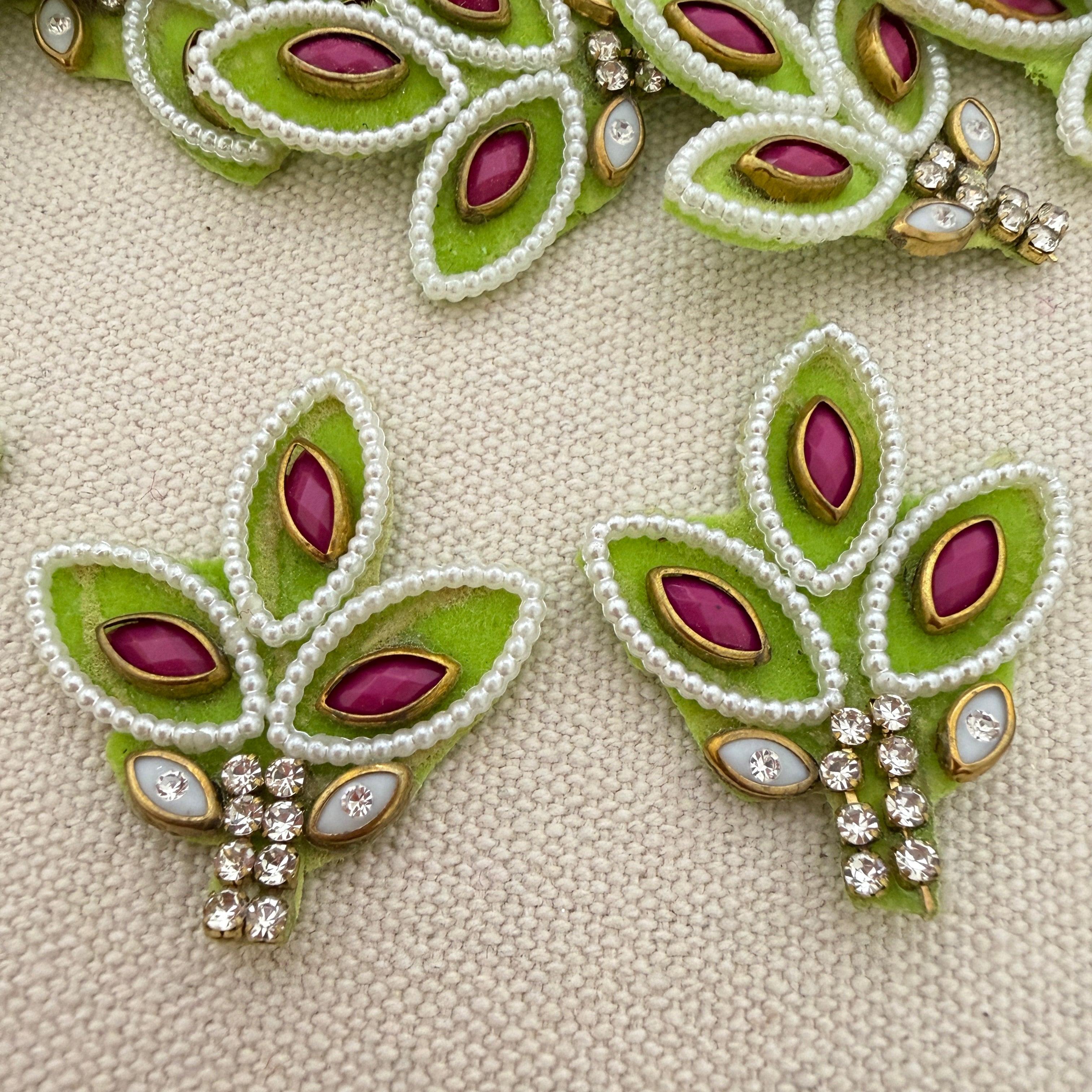 Decorative earrings with green leaves, purple stones, and white beads on a beige fabric background.