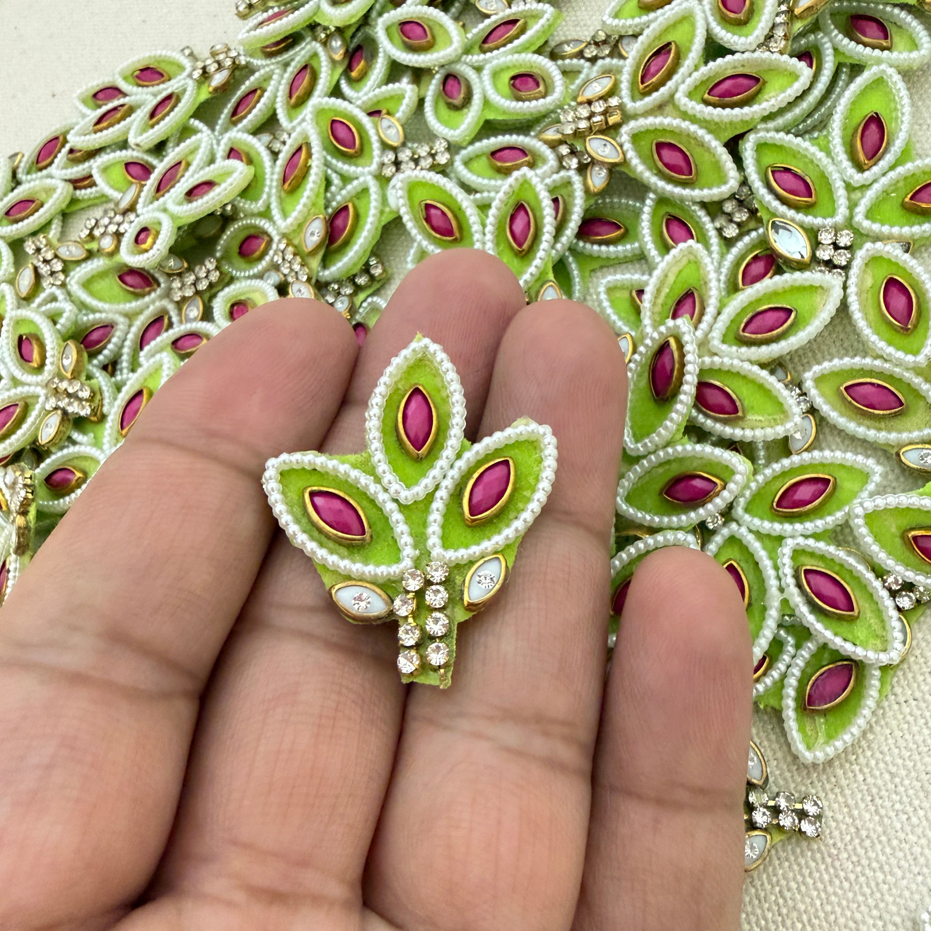 Decorative green and pink brooch held between fingers with a detailed embroidered background.